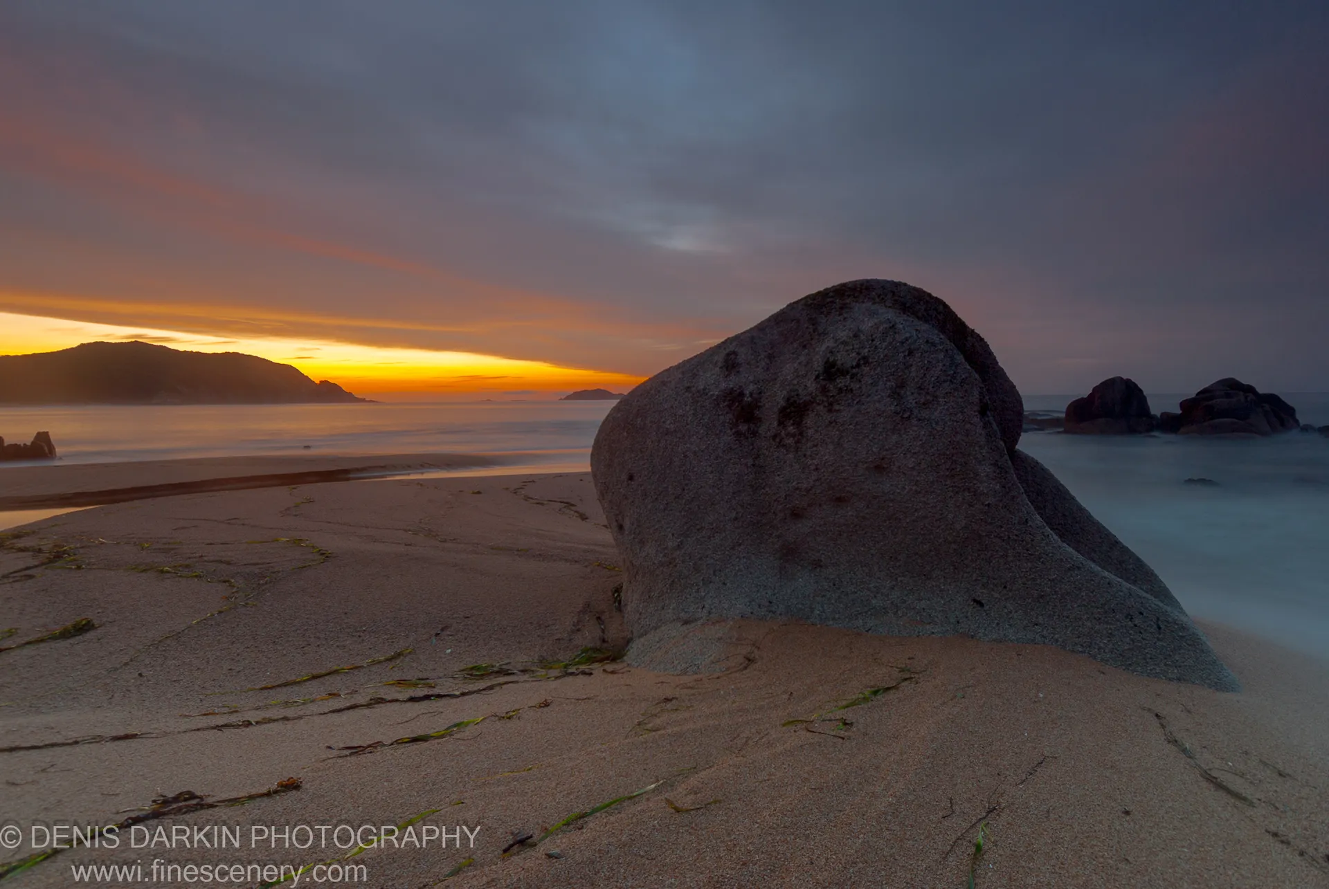 A large boulder on the sandy beach at Tachingouza Bay, with long-exposure surf as mist and a warm orange horizon at dawn.