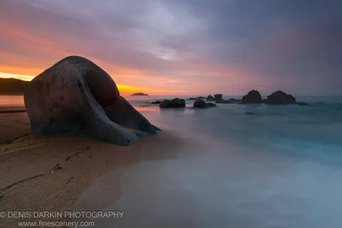 A massive weathered boulder on a reddish beach at Tachingouza Bay, with long-exposure surf flowing as mist around smaller rocks at dawn.