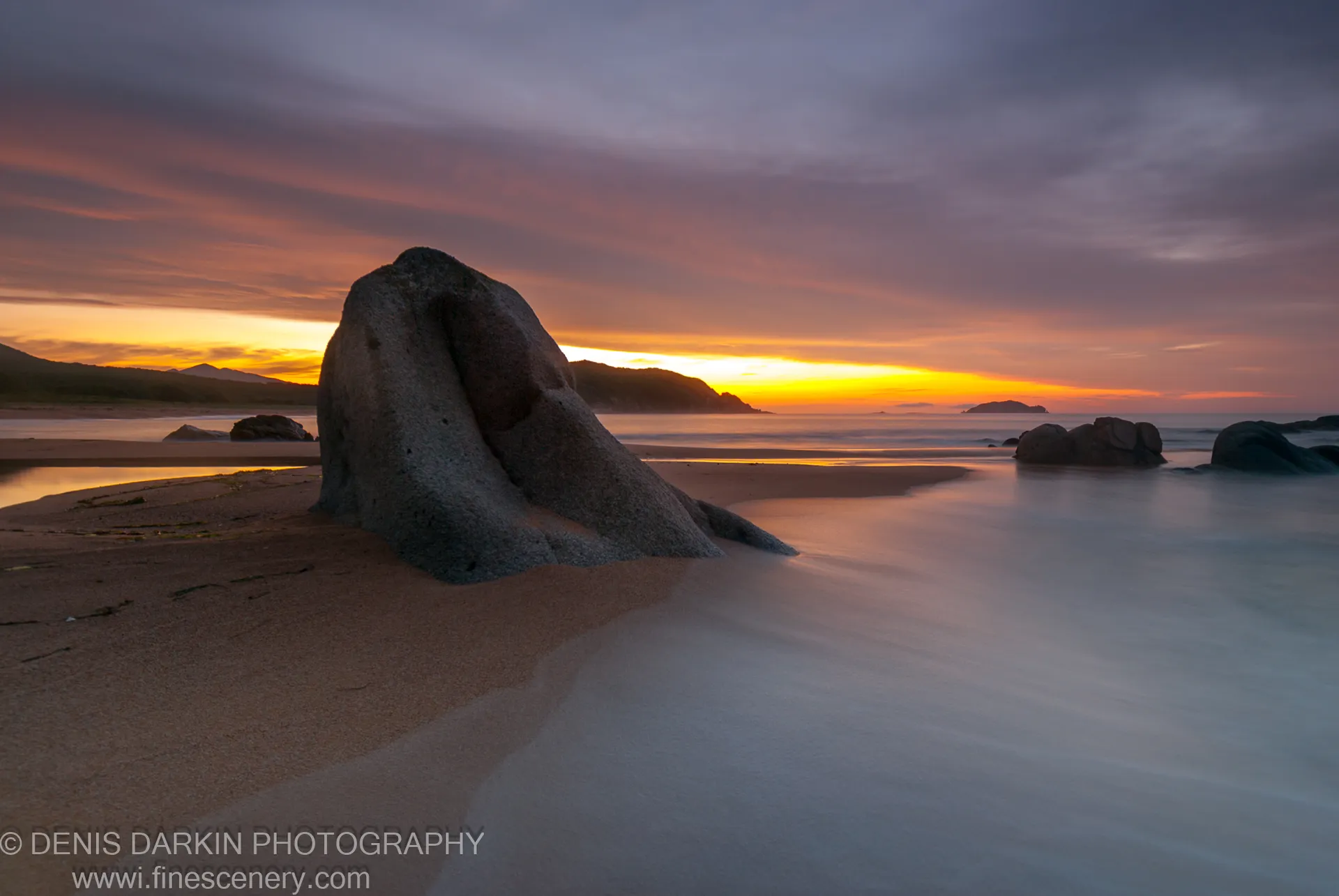 A massive smooth boulder leans into the sandy beach at Tachingouza Bay, long-exposure ocean beneath a violet and amber sky at dawn.