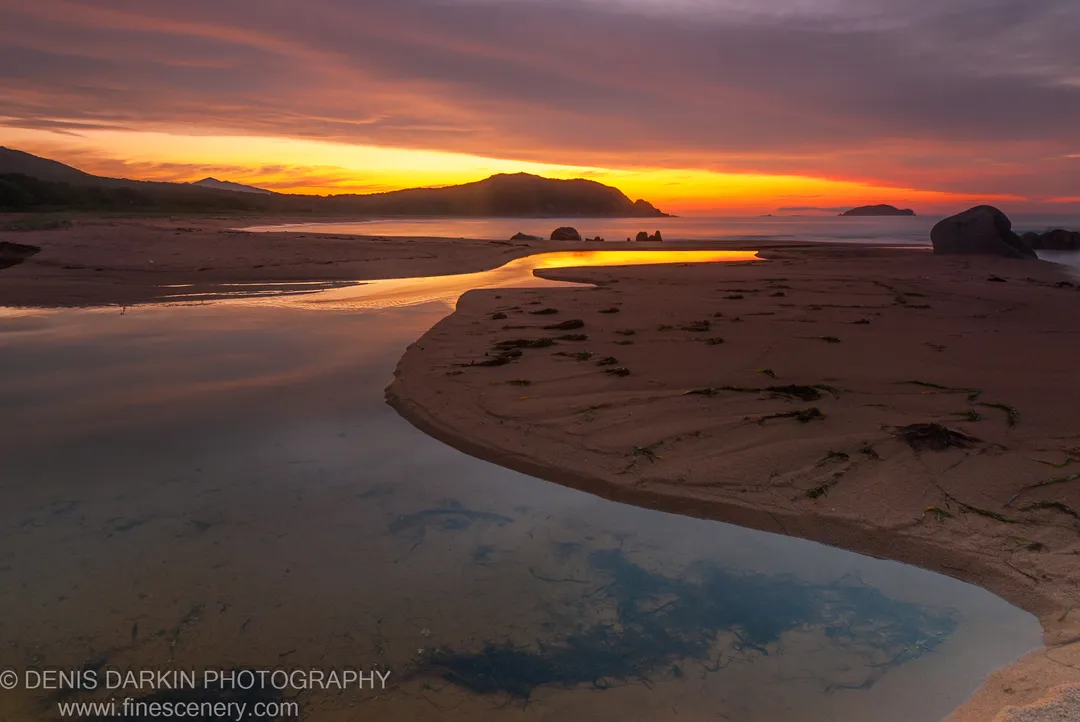 A winding tidal pool at Tachingouza Bay reflects an orange and purple dawn sky, with wet sand, scattered seaweed, and silhouetted headlands beyond.