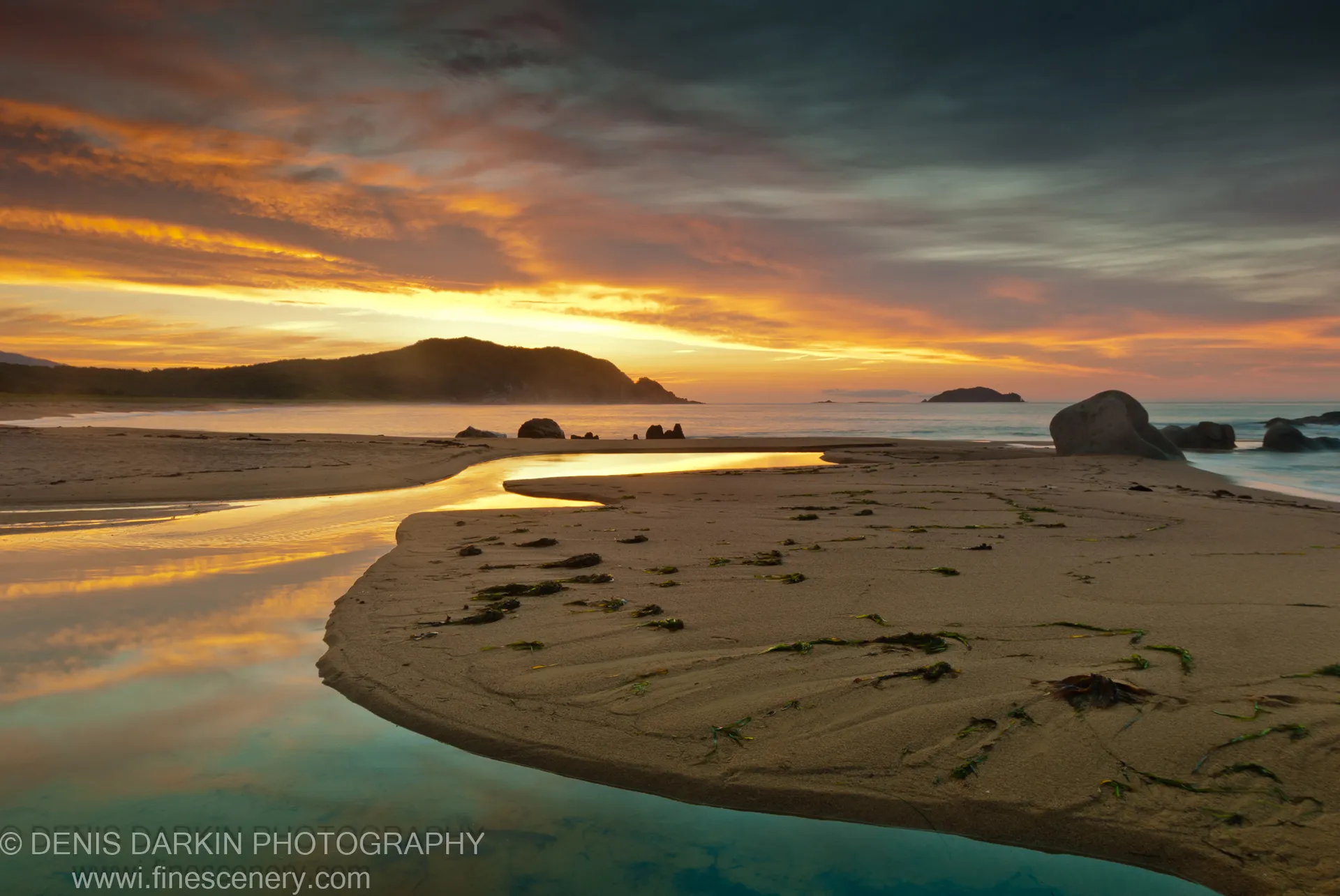 A winding pool at Tachingouza Bay reflects orange clouds in a dawn sky, with scattered seaweed, smooth rocks, and a silhouetted headland.