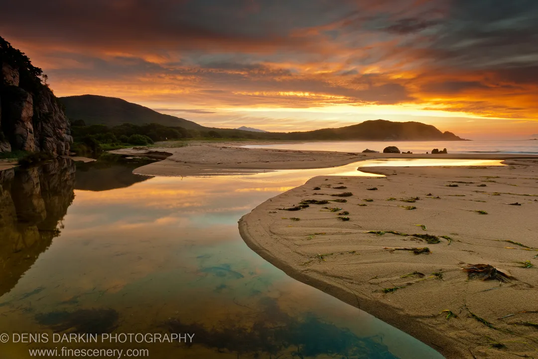 Stranded seaweed on wet sand at Tachingouza Bay at dawn, with a tidal stream winding toward dark rock cliffs under an orange sky.
