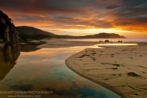 Stranded seaweed on wet sand at Tachingouza Bay at dawn, with a tidal stream winding toward dark rock cliffs under an orange sky.