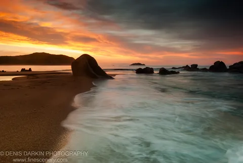 Long-exposure waves wash against a rounded granite boulder at Tachingouza Bay, with a silhouetted headland under an orange dawn sky.