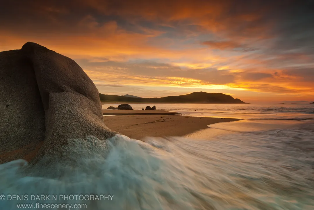 A massive smooth boulder in the left foreground at Tachingouza Bay at dawn, with blurred surf across wet sand and silhouetted hills on the horizon.