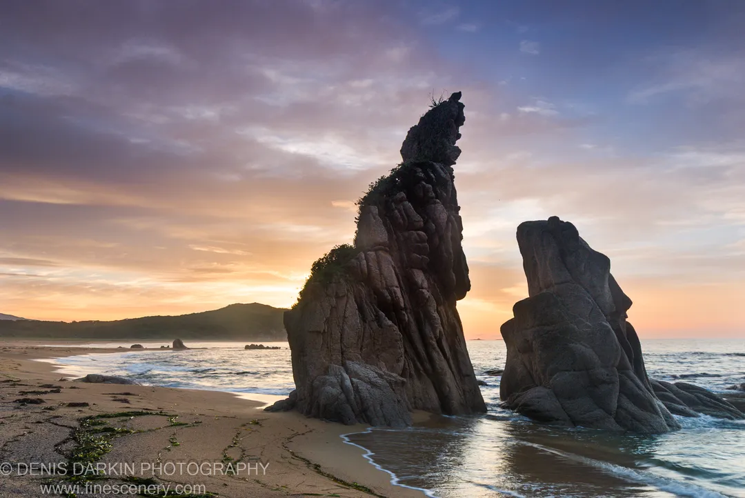 Towering granite sea stacks rising from the surf at Tachingouza Bay in morning light, with green seaweed and a sandy foreground below.