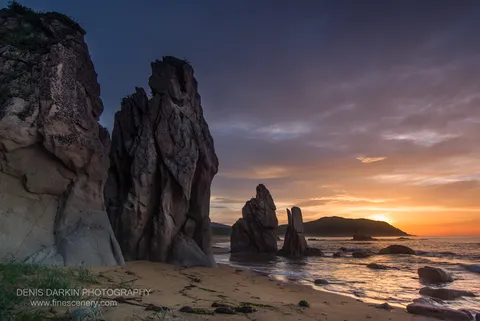 Massive weathered sea stacks in the left foreground at Tachingouza Bay at dawn, warm orange horizon above wet sand below.