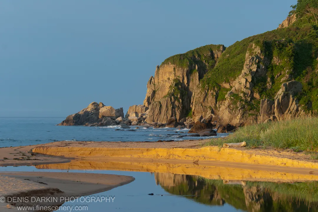 Morning sunlight on coastal cliffs and a sandy spit at Tachingouza Bay, with a calm tidal pool in the foreground.