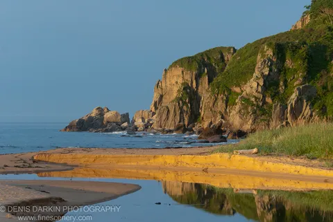 Morning sunlight on coastal cliffs and a sandy spit at Tachingouza Bay, with a calm tidal pool in the foreground.