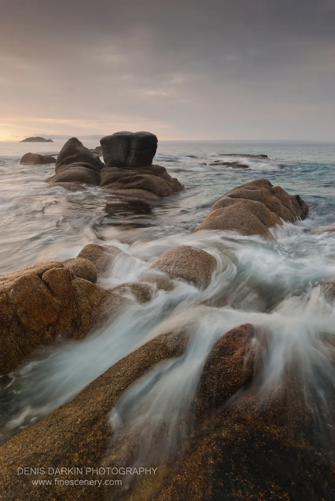 Rounded granite boulders rise from the sea near a distant islet at Tachingouza Bay, their surfaces softened by silky long-exposure water in the morning light.