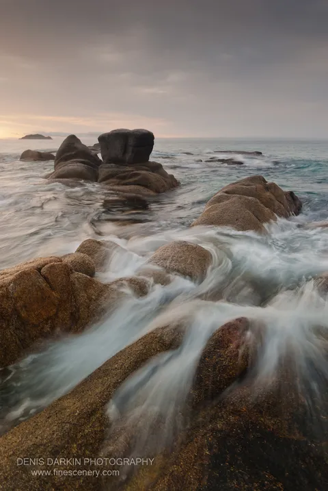 Rounded granite boulders rise from the sea near a distant islet at Tachingouza Bay, their surfaces softened by silky long-exposure water in the morning light.