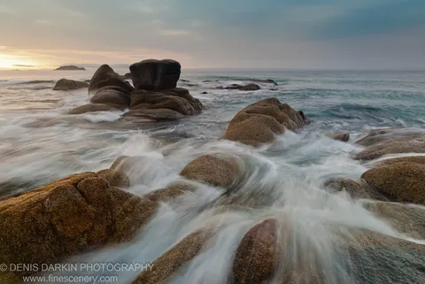 Warm morning sunlight catches the texture of granite rocks at Tachingouza Bay, where long-exposure water flows smoothly over foreground stone toward a distant island.