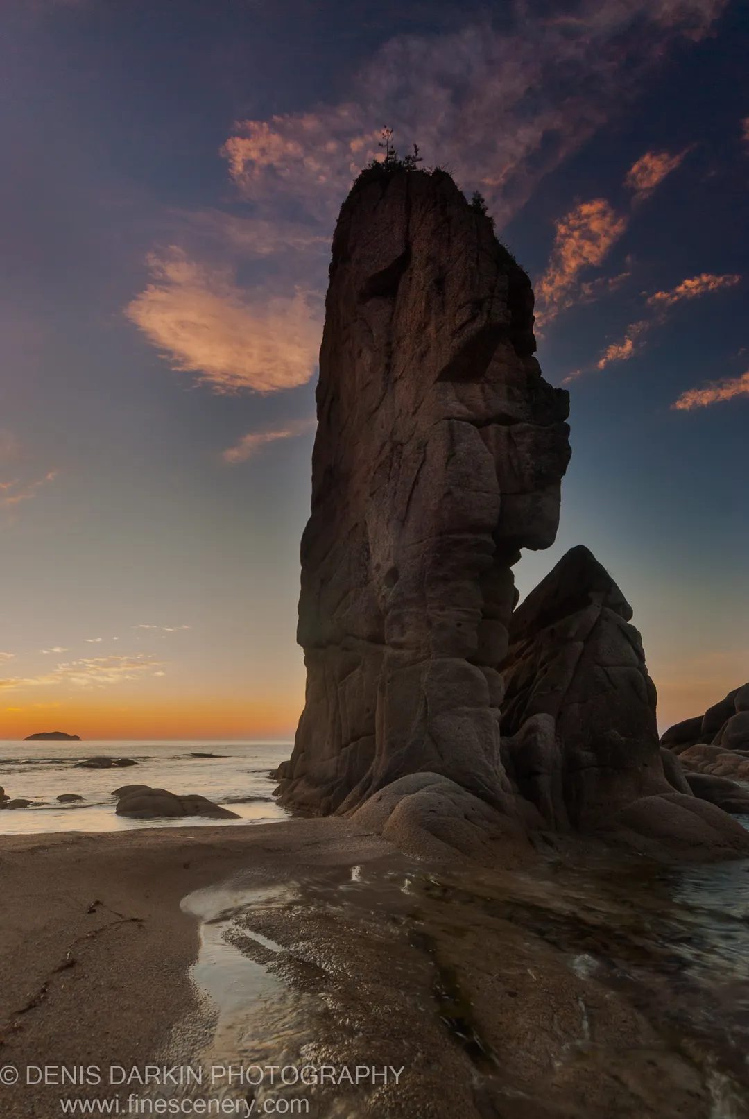 A massive rock formation rises from the Tachingouza Bay shoreline, its craggy face looking over wet foreground sand toward a distant island.