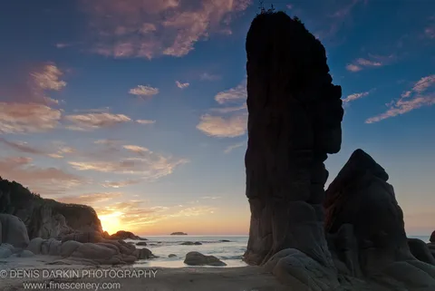A sea stack rises against the early morning sky at Tachingouza Bay, with smooth rounded boulders and wet sand in the foreground.