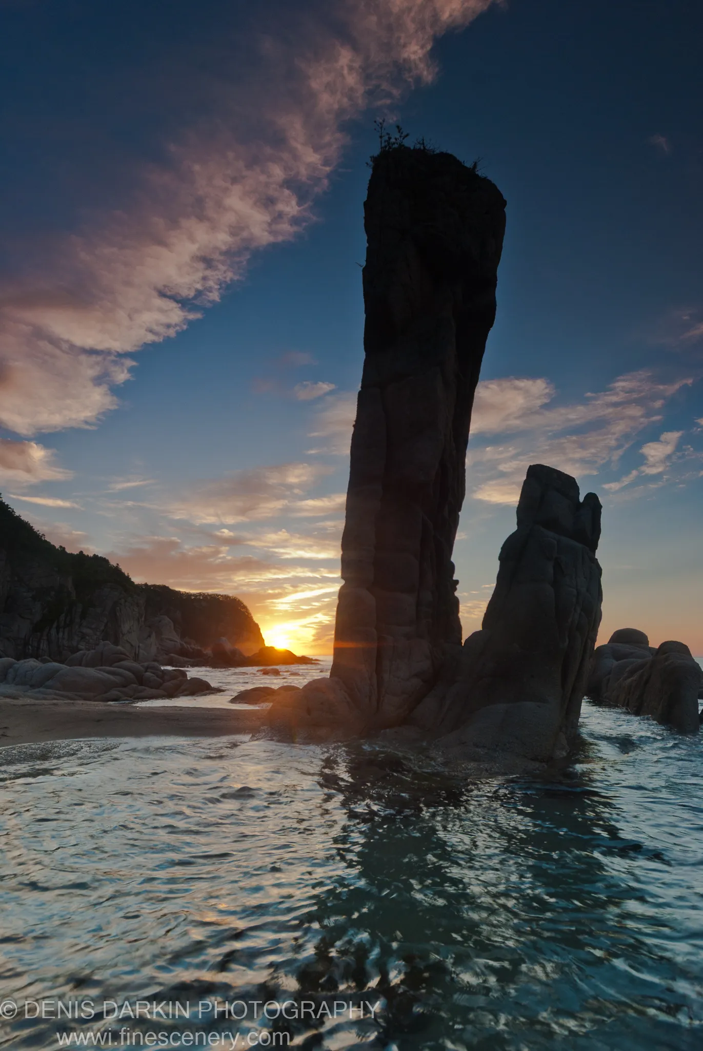 A sea stack in dark silhouette against the morning sky at Tachingouza Bay, with rippling water catching the light below.