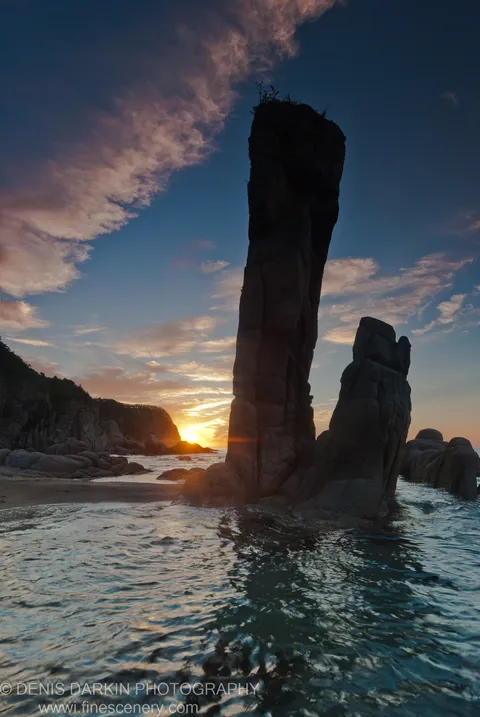 A sea stack in dark silhouette against the morning sky at Tachingouza Bay, with rippling water catching the light below.