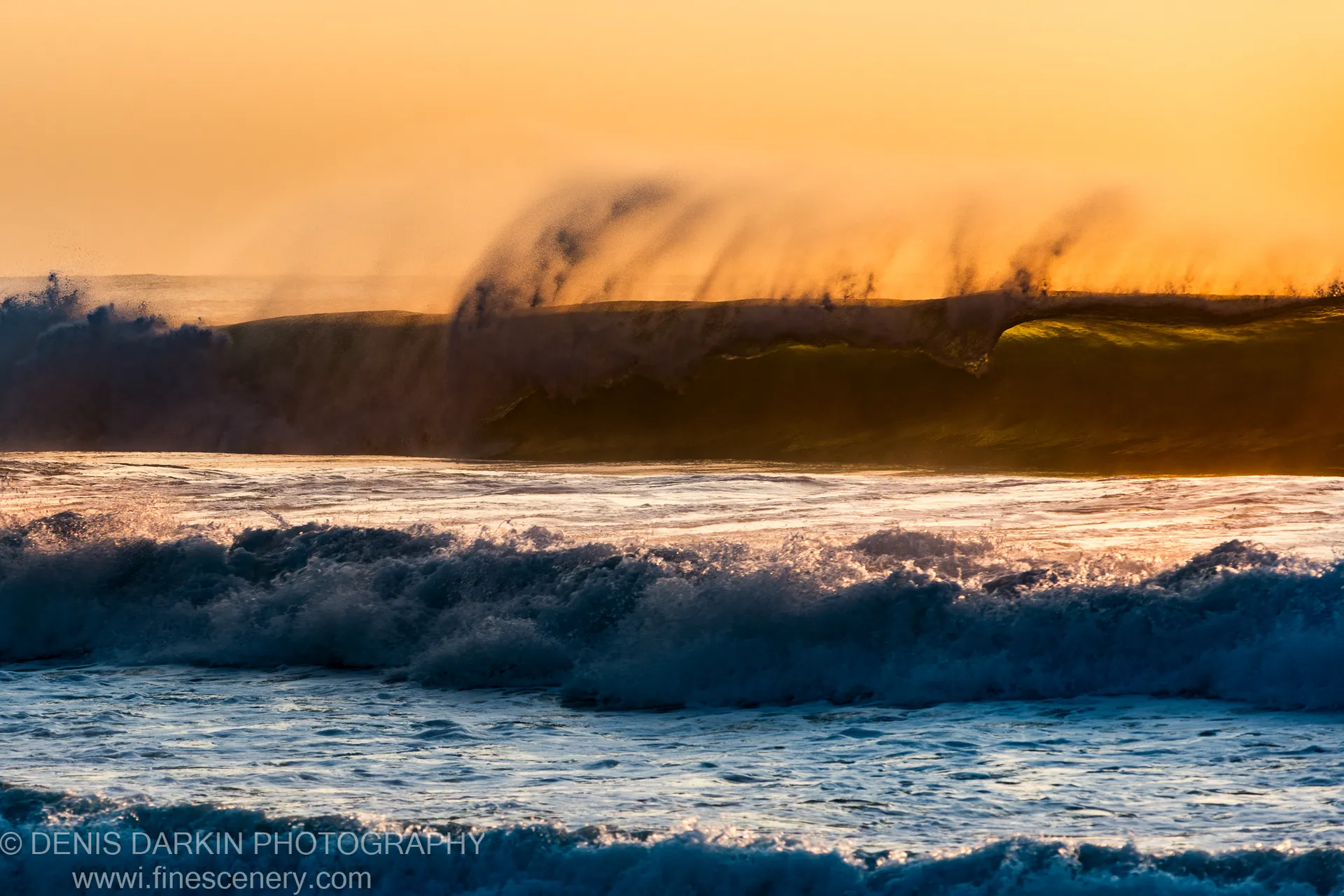 A large wave crests and sends a plume of mist into the air against an orange-lit dawn sky at Spoon Bay.