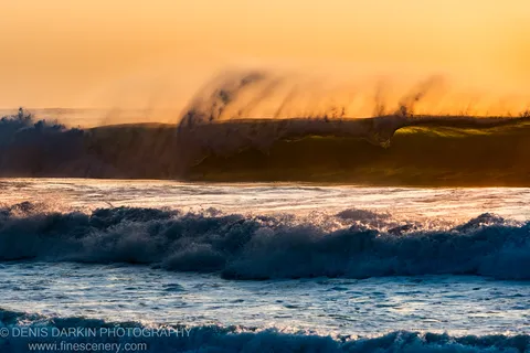 A large wave crests and sends a plume of mist into the air against an orange-lit dawn sky at Spoon Bay.