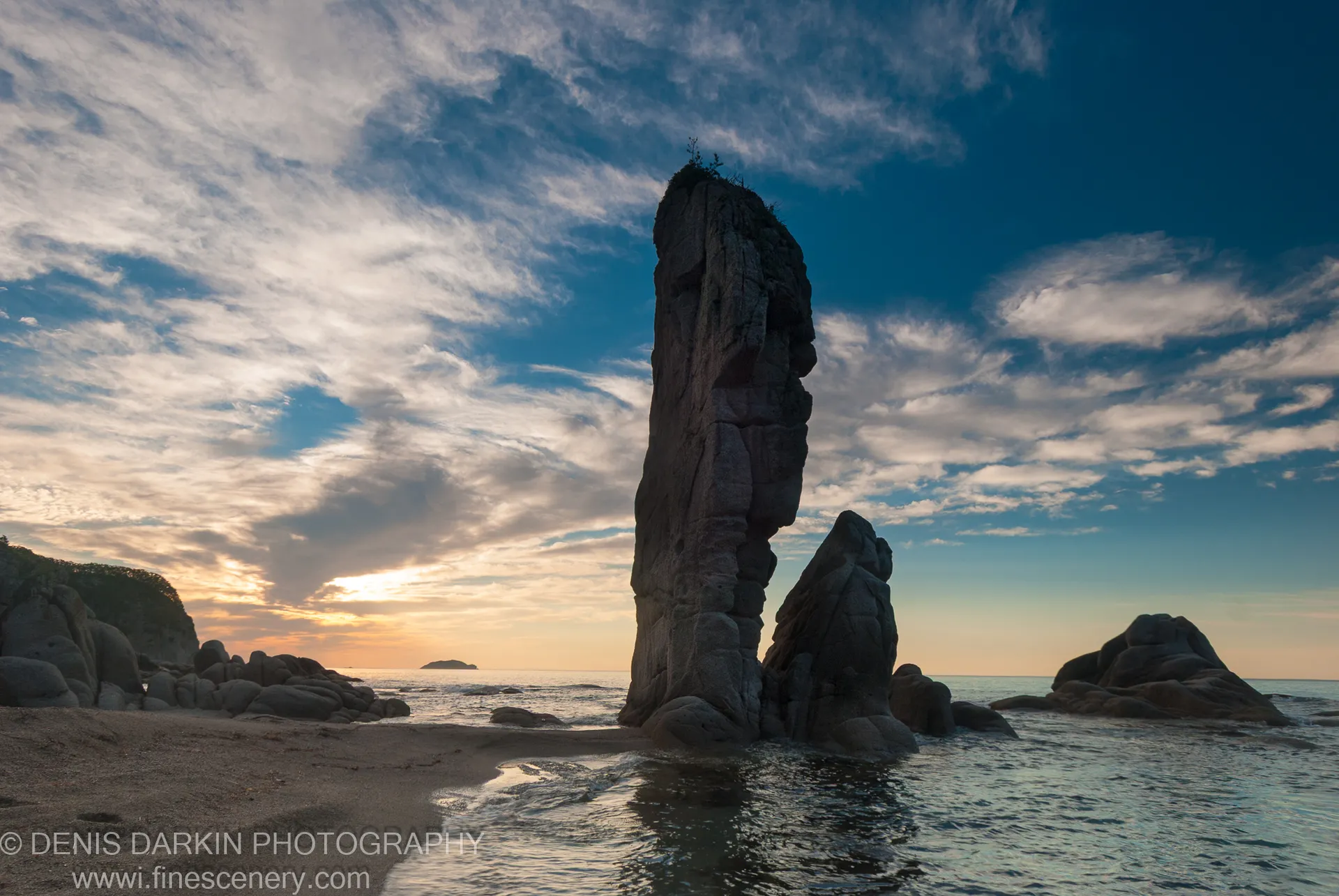 A massive sea stack with a rough rock face dominates the foreground at Tachingouza Bay, with morning light spreading across the ocean and sand below.