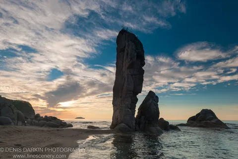 A massive sea stack with a rough rock face dominates the foreground at Tachingouza Bay, with morning light spreading across the ocean and sand below.