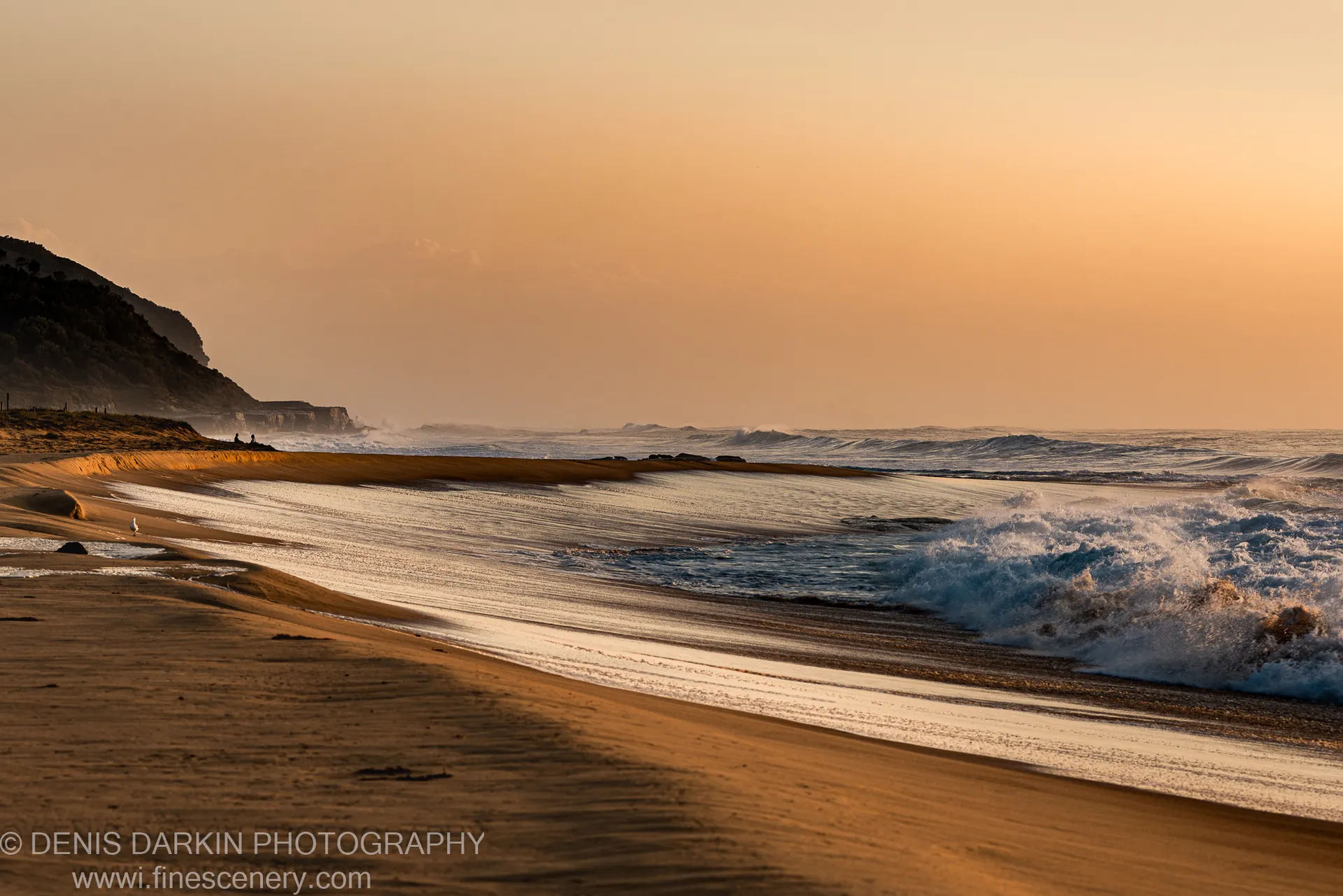 Wet sand stretches along the Spoon Bay shoreline toward breaking surf and the silhouetted headland in warm amber morning light.