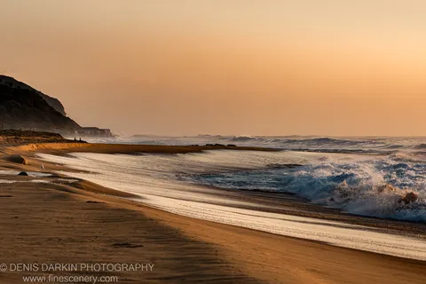 Wet sand stretches along the Spoon Bay shoreline toward breaking surf and the silhouetted headland in warm amber morning light.