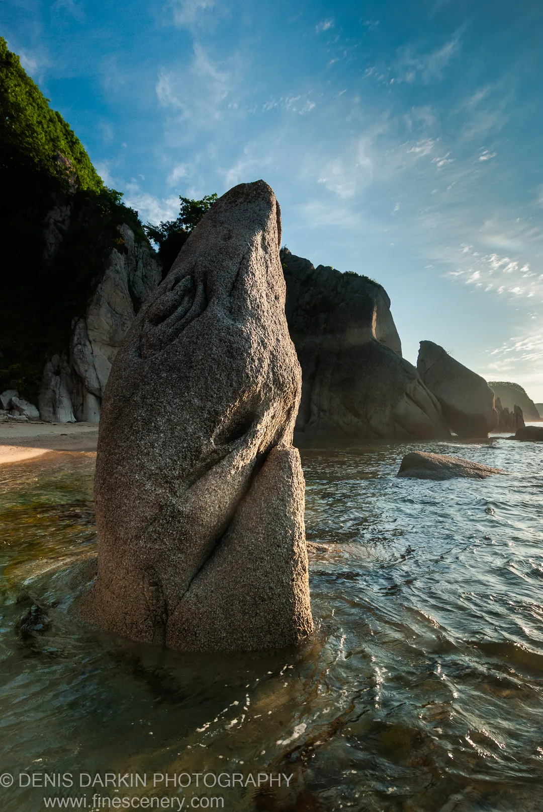 A tapered granite monolith rises from the surf at Tachingouza Bay, its rough surface catching low morning sun with mossy cliffs and cirrus sky behind.