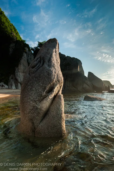 A tapered granite monolith rises from the surf at Tachingouza Bay, its rough surface catching low morning sun with mossy cliffs and cirrus sky behind.