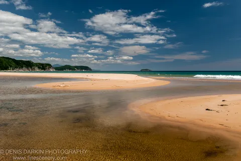 A sandbar arcs through sun-lit shallow water at Tachingouza Bay, with green cliffs on the left and white cumulus clouds overhead.