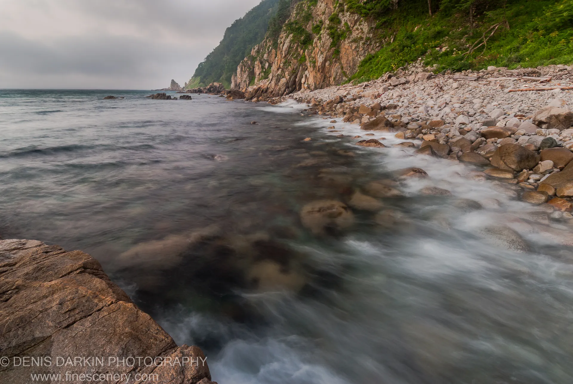 Silky long-exposure water flows over pebbles and boulders toward tree-draped cliffs at Tachingouza Bay in the pre-dawn light of August.