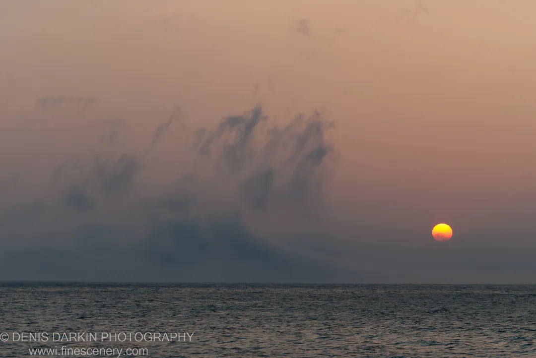 A bright morning sun sits low over dark rippled water at Tachingouza Bay, with textured clouds to one side against the haze.