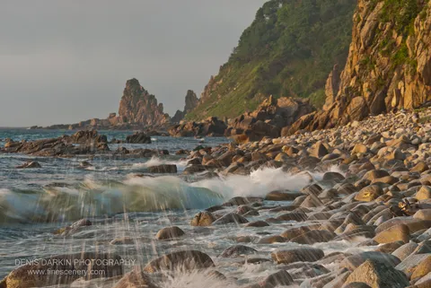 Warm morning sun on a vegetated coastal cliff at Tachingouza Bay, with smooth waves over foreground stones and sea stacks in the distance.