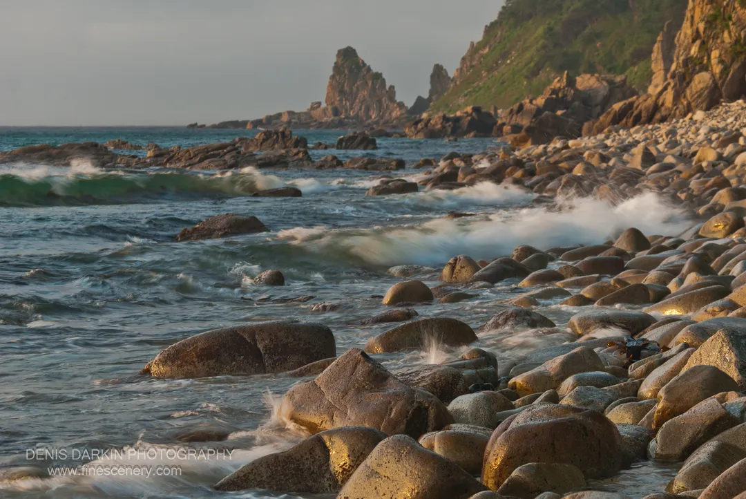 Smooth boulders in morning sunlight at Tachingouza Bay, with white surf in the foreground and jagged rock spires rising in the distance.