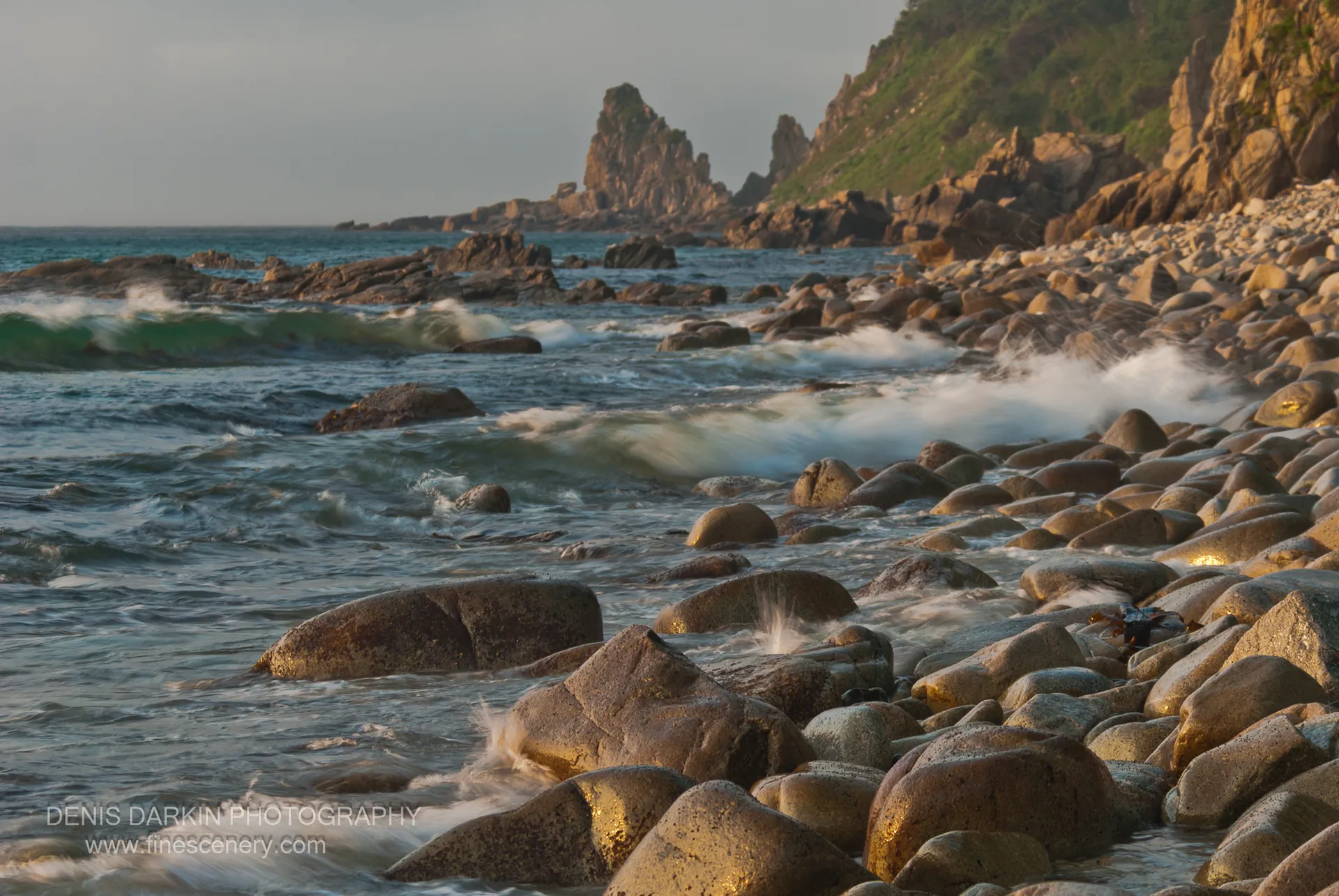 Smooth boulders in morning sunlight at Tachingouza Bay, with white surf in the foreground and jagged rock spires rising in the distance.