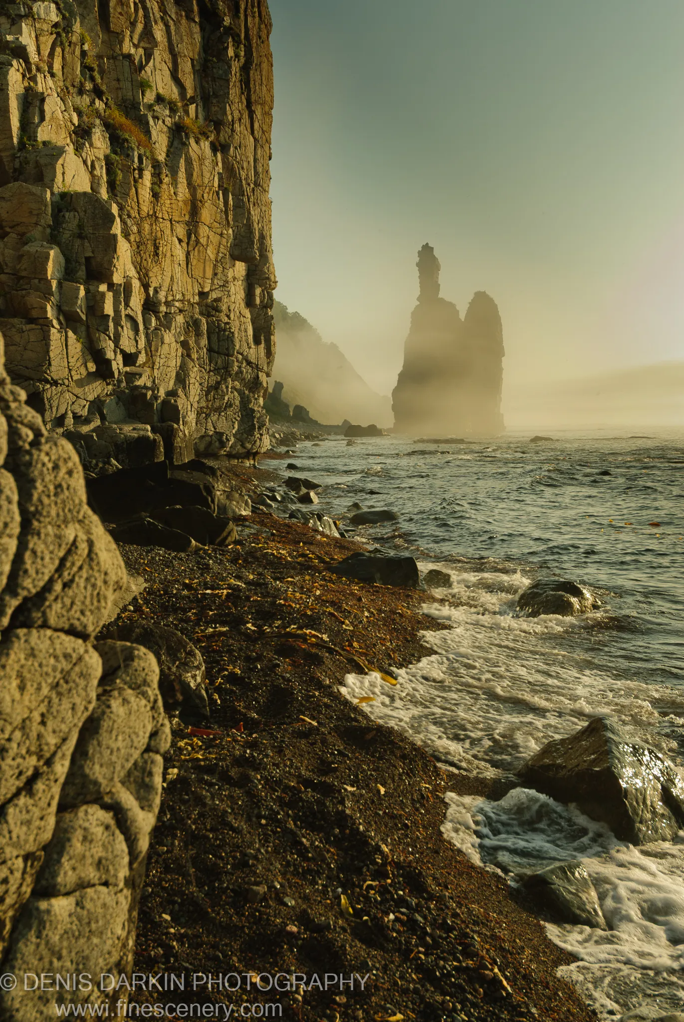 A dark shingle beach strewn with seaweed stretches toward granite sea stacks emerging from dense morning mist at Dalnegorsk, Primorsky Krai.