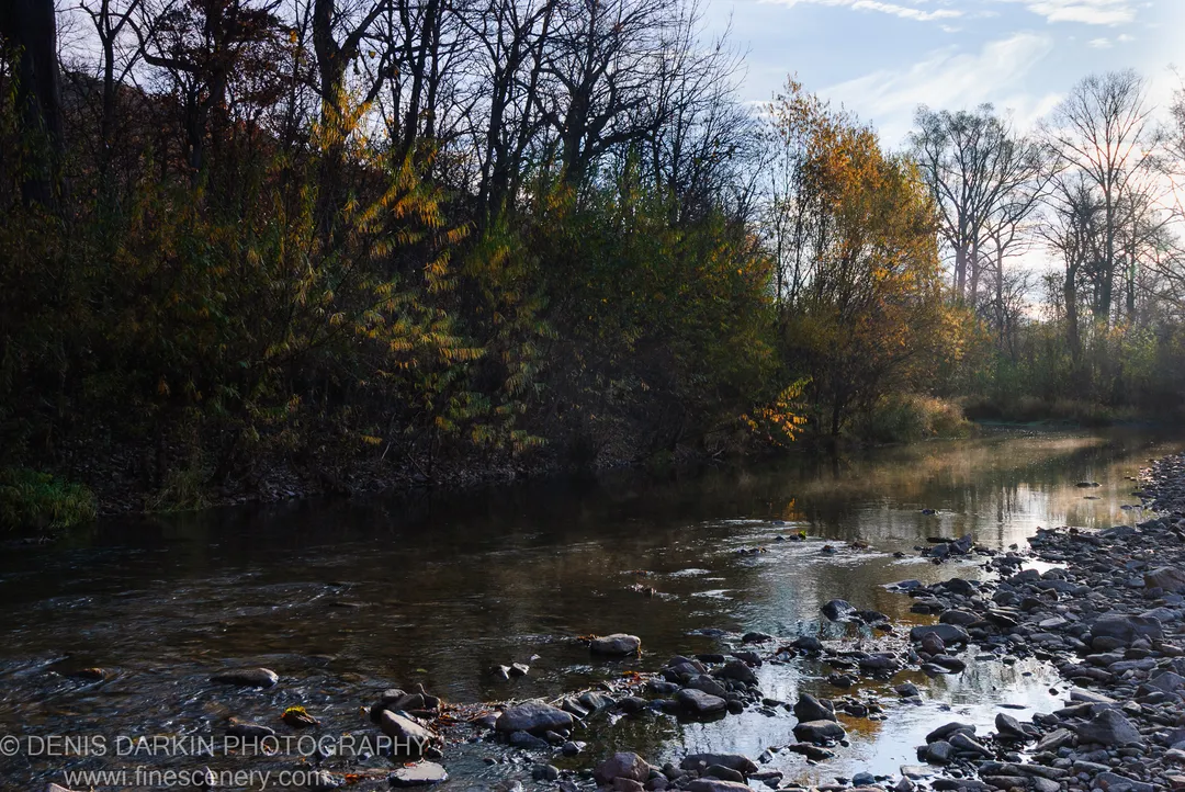 A river meanders past bare-branched trees at the edge of a rocky bank, with morning sunlight on the water and autumnal foliage in the background.