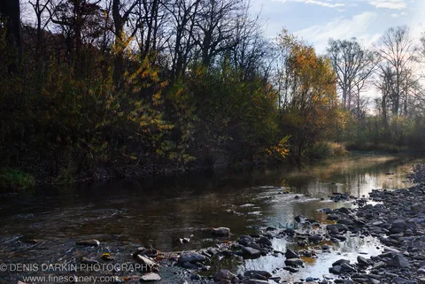 A river meanders past bare-branched trees at the edge of a rocky bank, with morning sunlight on the water and autumnal foliage in the background.
