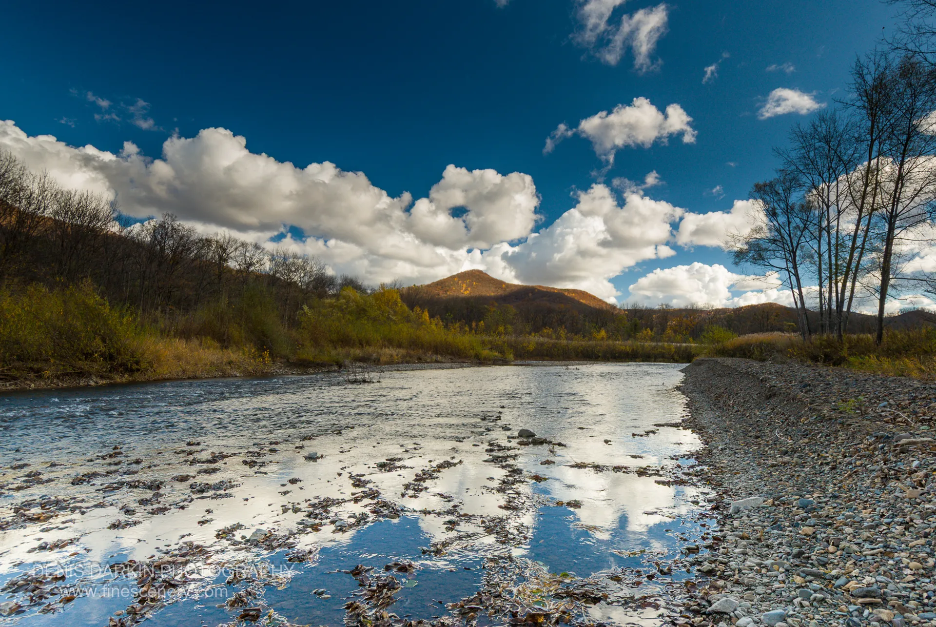 A wide, shallow river with a rocky bank reflects a cloud-filled sky, golden autumn foliage covering the distant hills, Daubihe River, Primorsky Krai.