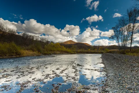 A wide, shallow river with a rocky bank reflects a cloud-filled sky, golden autumn foliage covering the distant hills, Daubihe River, Primorsky Krai.