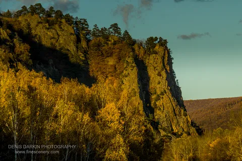 A sheer rock face lit by afternoon sun, yellow foliage at its base and dark pines on the summit, Lefu River valley, Primorsky Krai.