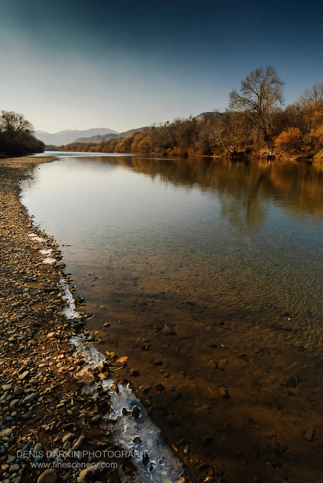 Warm sunlight catches a thin ridge of ice along the Suchan riverbank, with submerged stones visible in the water and distant hazy hills beyond.