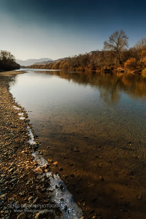 Warm sunlight catches a thin ridge of ice along the Suchan riverbank, with submerged stones visible in the water and distant hazy hills beyond.