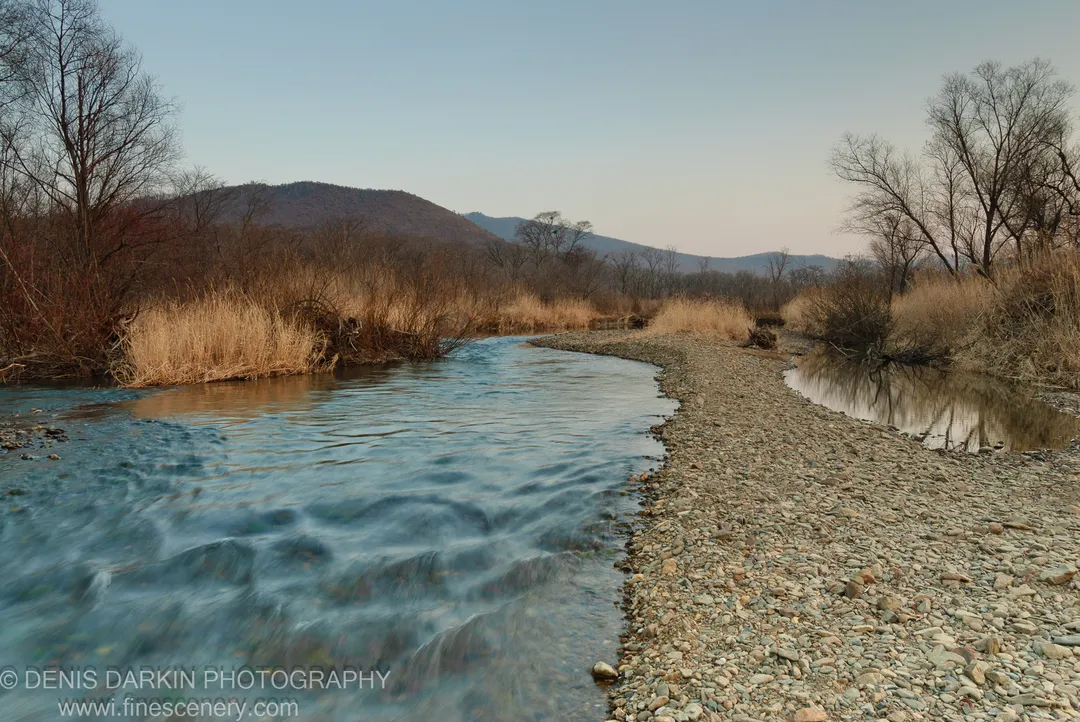 The Komissarovka River flows through a wide valley past dry reeds and bare trees, with distant mountains visible at dusk.