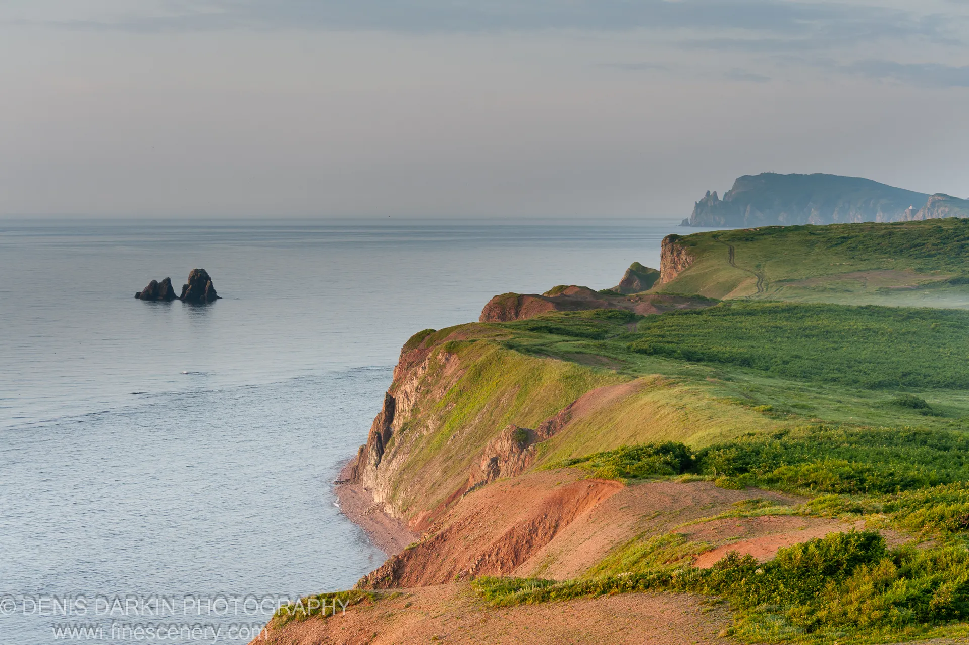Verdant clifftop slopes fall sharply to eroded red clay cliffs and a calm slate-grey sea on the Vladimir Bay coast in summer.
