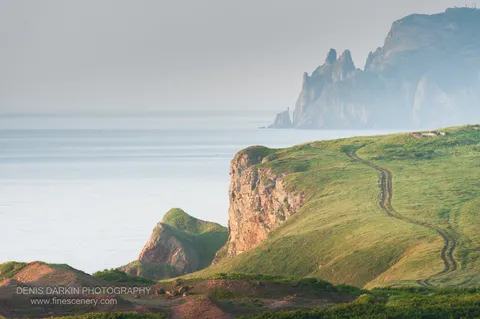 A winding dirt track follows the grassy headland edge at Vladimir Bay, warm light on the cliff face and a pale sea beyond.