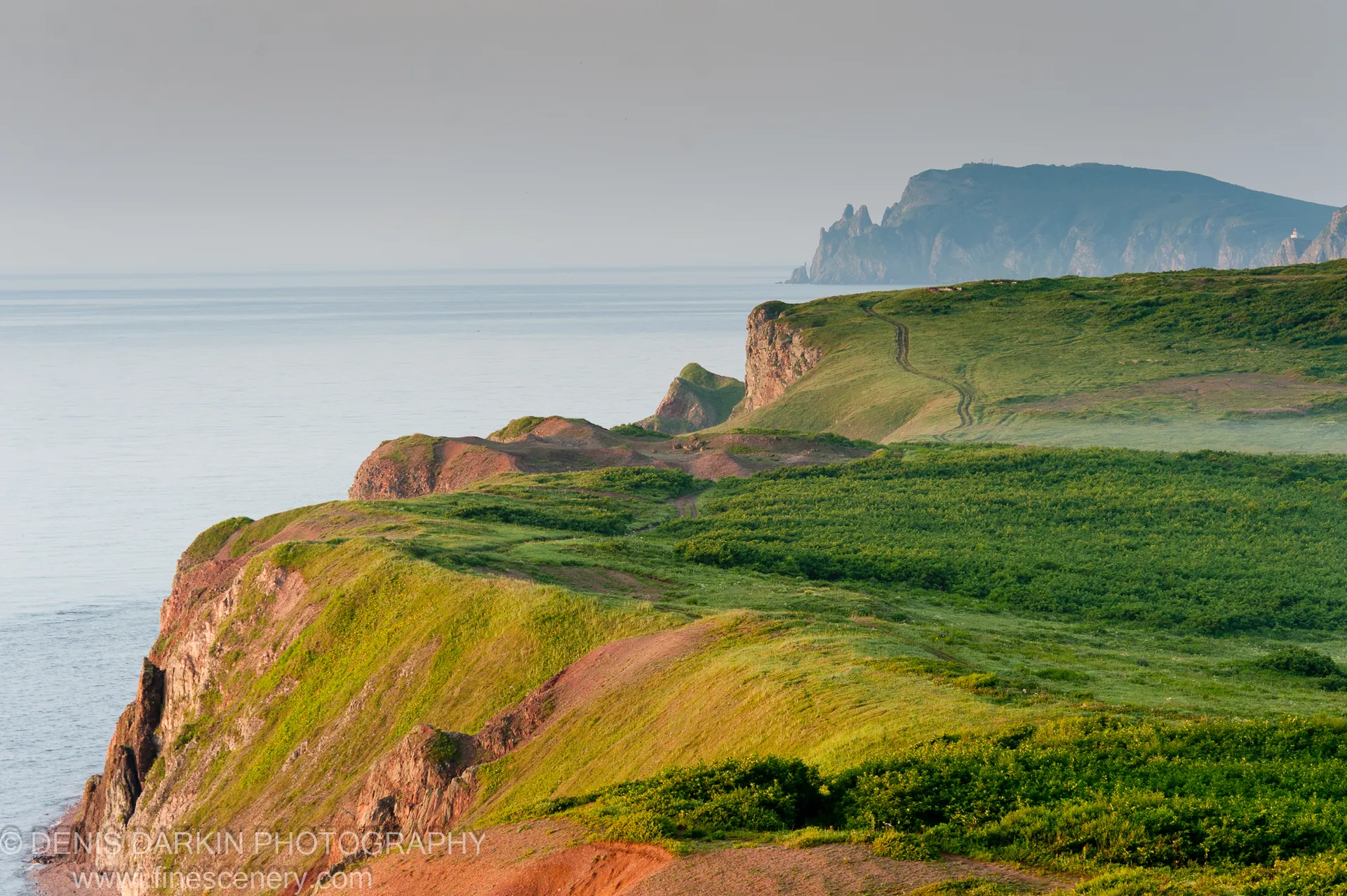 Lush green slopes give way to red-soil cliffs and rocky pinnacles along the Vladimir Bay coast, with a winding track following the ridgeline.