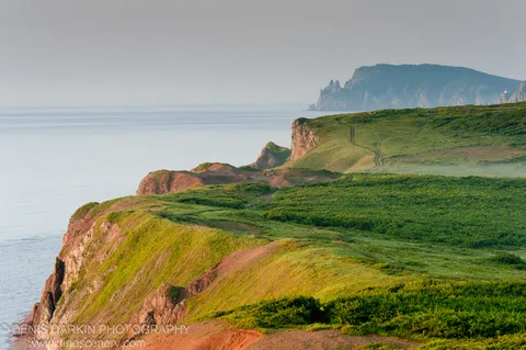 Lush green slopes give way to red-soil cliffs and rocky pinnacles along the Vladimir Bay coast, with a winding track following the ridgeline.