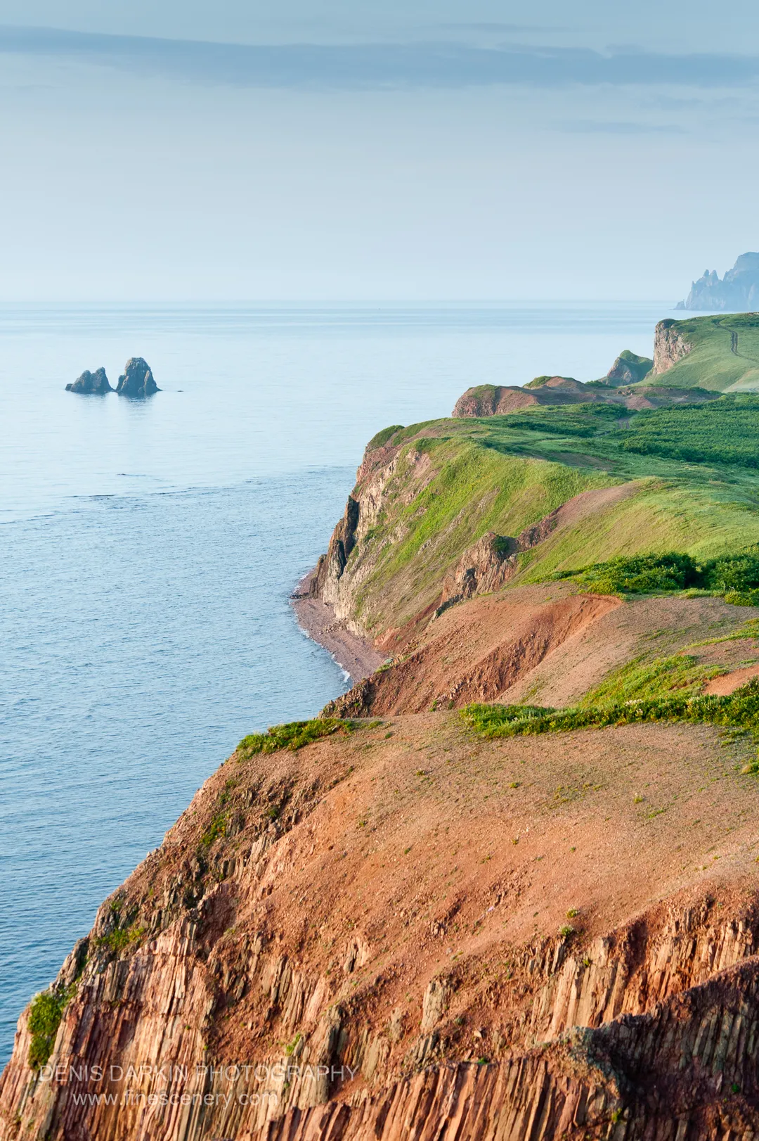 Columnar basalt formations rise from the cliff base along Vladimir Bay, reddish earth in the foreground and distant rock islets visible beyond.