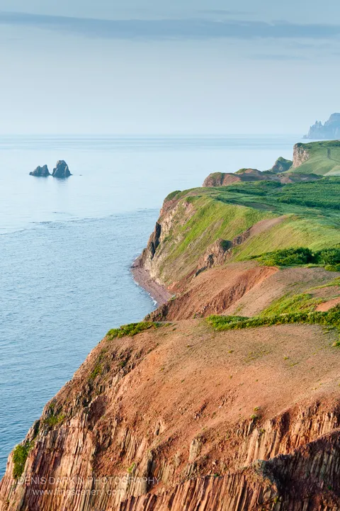 Columnar basalt formations rise from the cliff base along Vladimir Bay, reddish earth in the foreground and distant rock islets visible beyond.