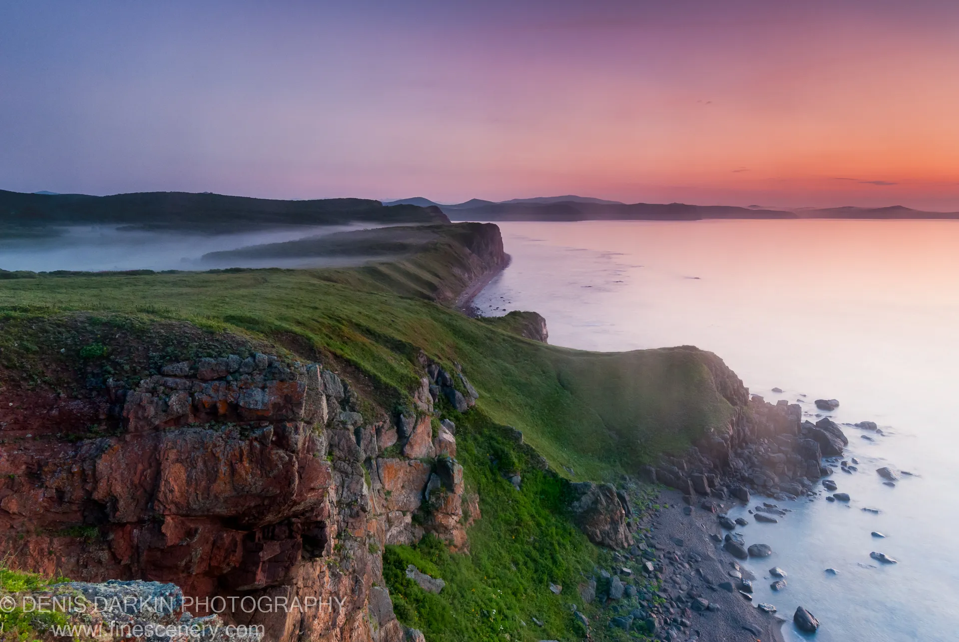 Reddish-brown cliff edge with mossy rocks and green grass overlooks a calm sea at dawn, Vladimir Bay, Primorsky Krai, Russia.